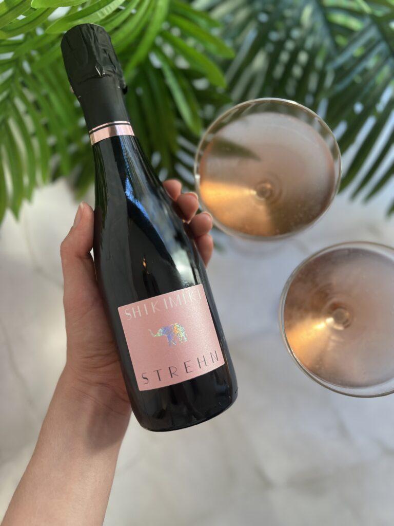Eine Flasche SHIKI MIKI Rosé Sekt und zwei elegante Gläser auf einem hellen Untergrund mit Pflanzen im Hintergrund.