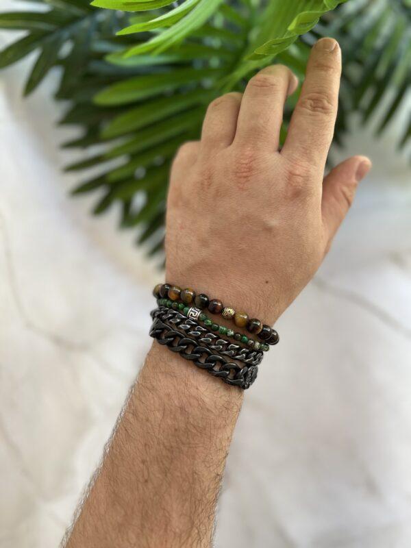 Männliche Hand mit mehreren modischen Armbändern, umgeben von tropischen Pflanzenblättern.